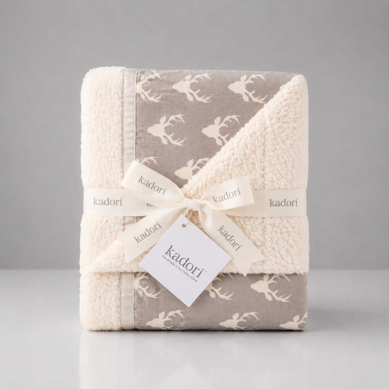 A folded Kadori baby blanket with a soft sherpa fleece texture, featuring a taupe-gray and cream deer pattern, wrapped in a cream ribbon with a Kadori tag.