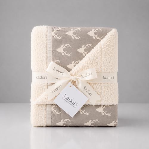 A folded Kadori baby blanket with a soft sherpa fleece texture, featuring a taupe-gray and cream deer pattern, wrapped in a cream ribbon with a Kadori tag.