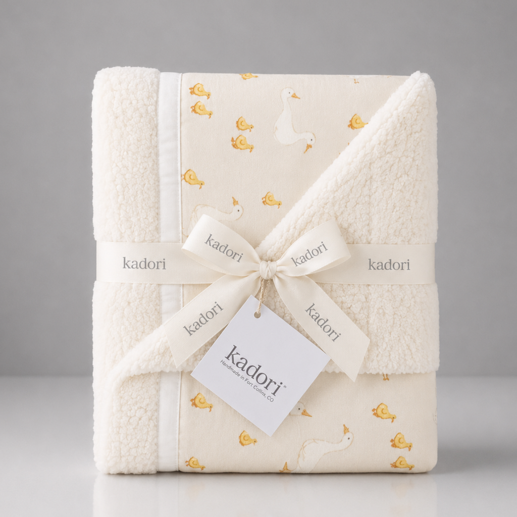 A folded Kadori baby blanket with a sherpa fleece border and a printed cream fabric center featuring a white goose and small yellow duck motifs. The blanket is wrapped in a cream ribbon printed with the Kadori brand name and tied with a white paper tag.