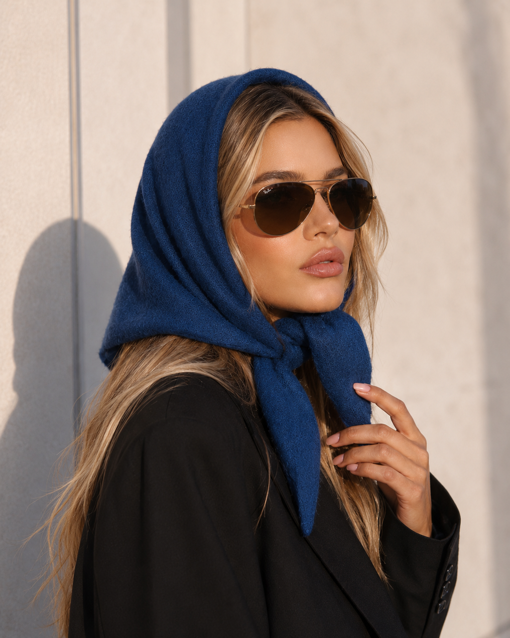 A woman wearing a deep blue knitted headscarf and dark sunglasses, posing in a black blazer against a textured light beige wall.