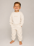 A smiling young boy stands barefoot against a light gray studio background, wearing a cream-colored sweatshirt and matching sweatpants with 'CSU' embroidered on the chest.