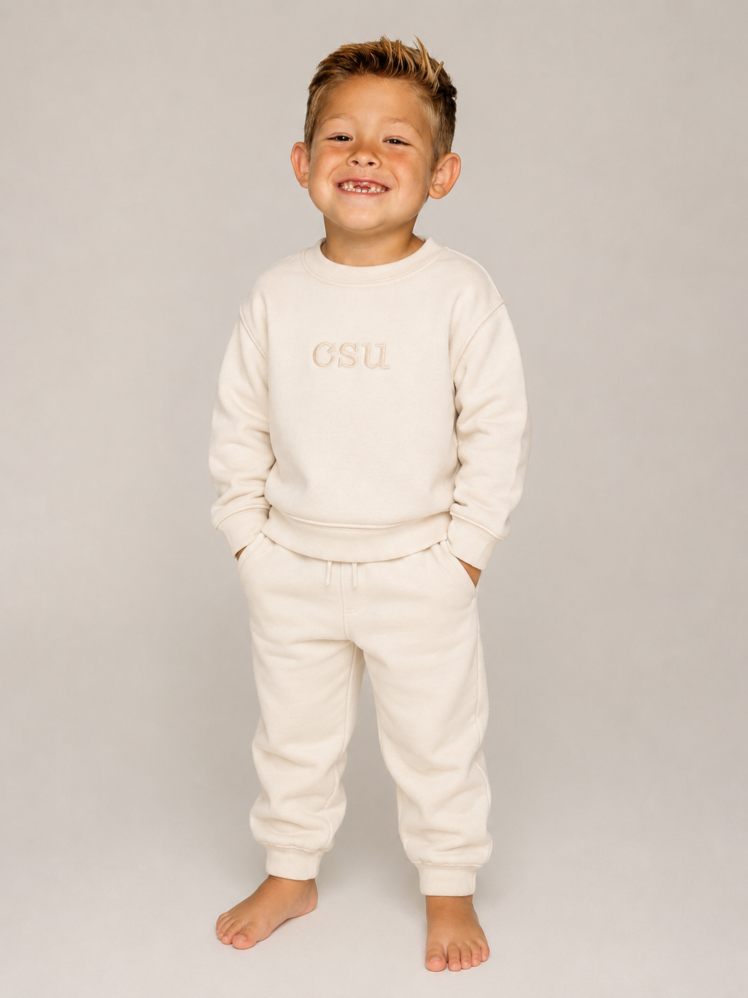 A smiling young boy stands barefoot against a light gray studio background, wearing a cream-colored sweatshirt and matching sweatpants with 'CSU' embroidered on the chest.
