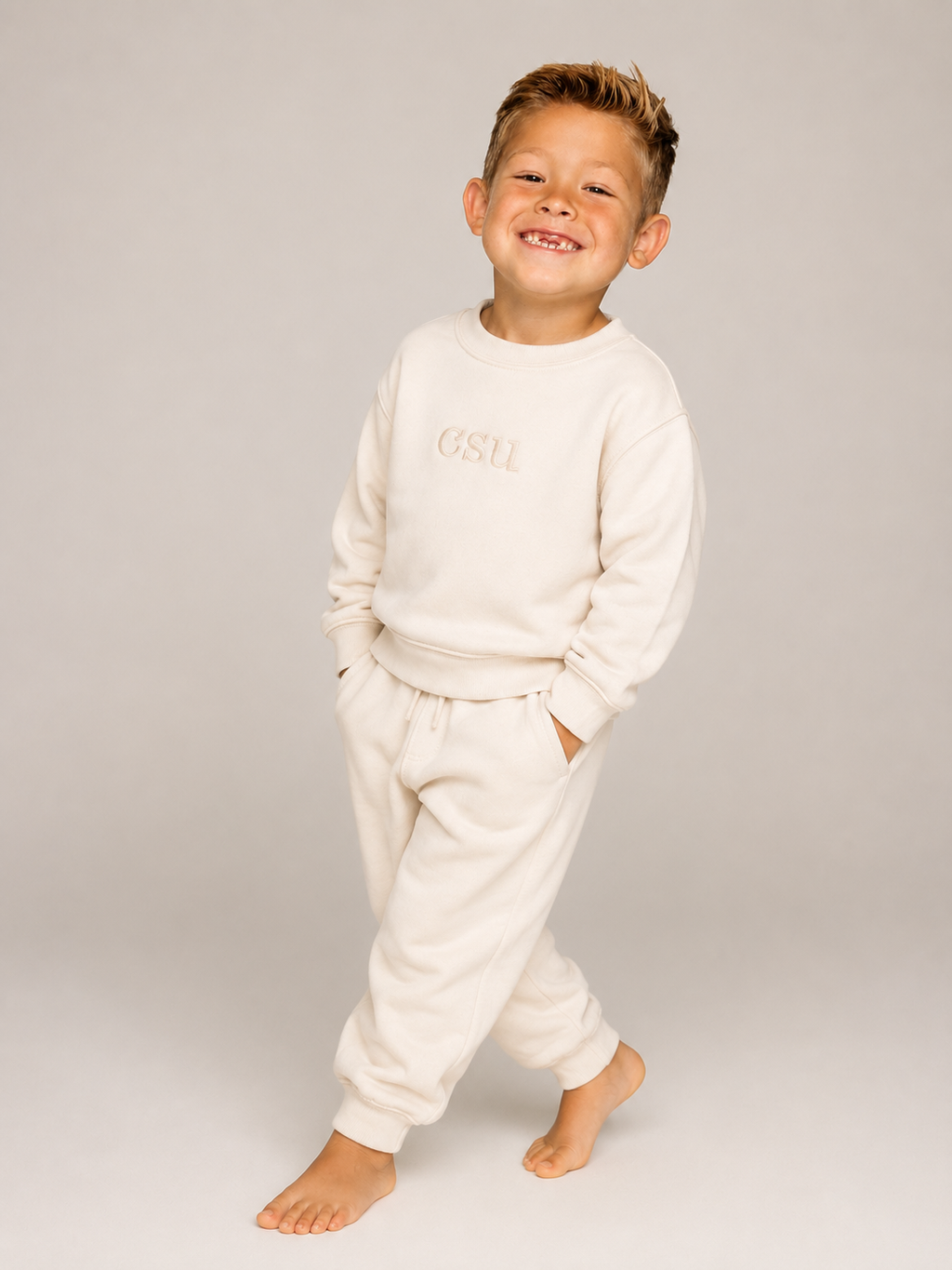 A young boy smiling and walking barefoot while wearing a cream-colored two-piece sweatshirt and jogger pants set with 'OSU' printed on the chest.
