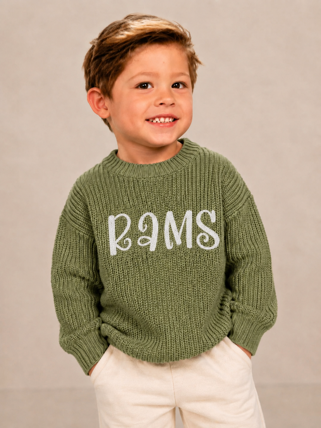 A young boy smiling and wearing an olive green ribbed knit sweater with 'RAMS' written in white across the chest, paired with cream pants, against a neutral gray studio background.