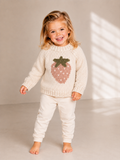 A young girl smiling and posing in a cream-colored knit sweater and matching sweatpants set. The sweater features a large, knitted pink strawberry design on the front with a green leafy top.
