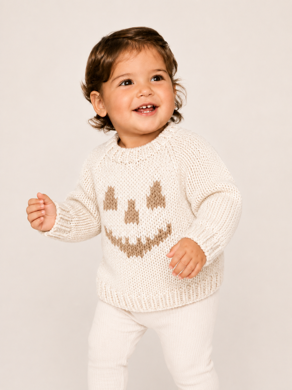 A smiling toddler wearing a cream knit sweater with a brown knitted jack-o'-lantern face and white pants, standing against a plain light beige studio background.