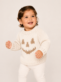 A smiling toddler wearing a cream knit sweater with a brown knitted jack-o'-lantern face and white pants, standing against a plain light beige studio background.