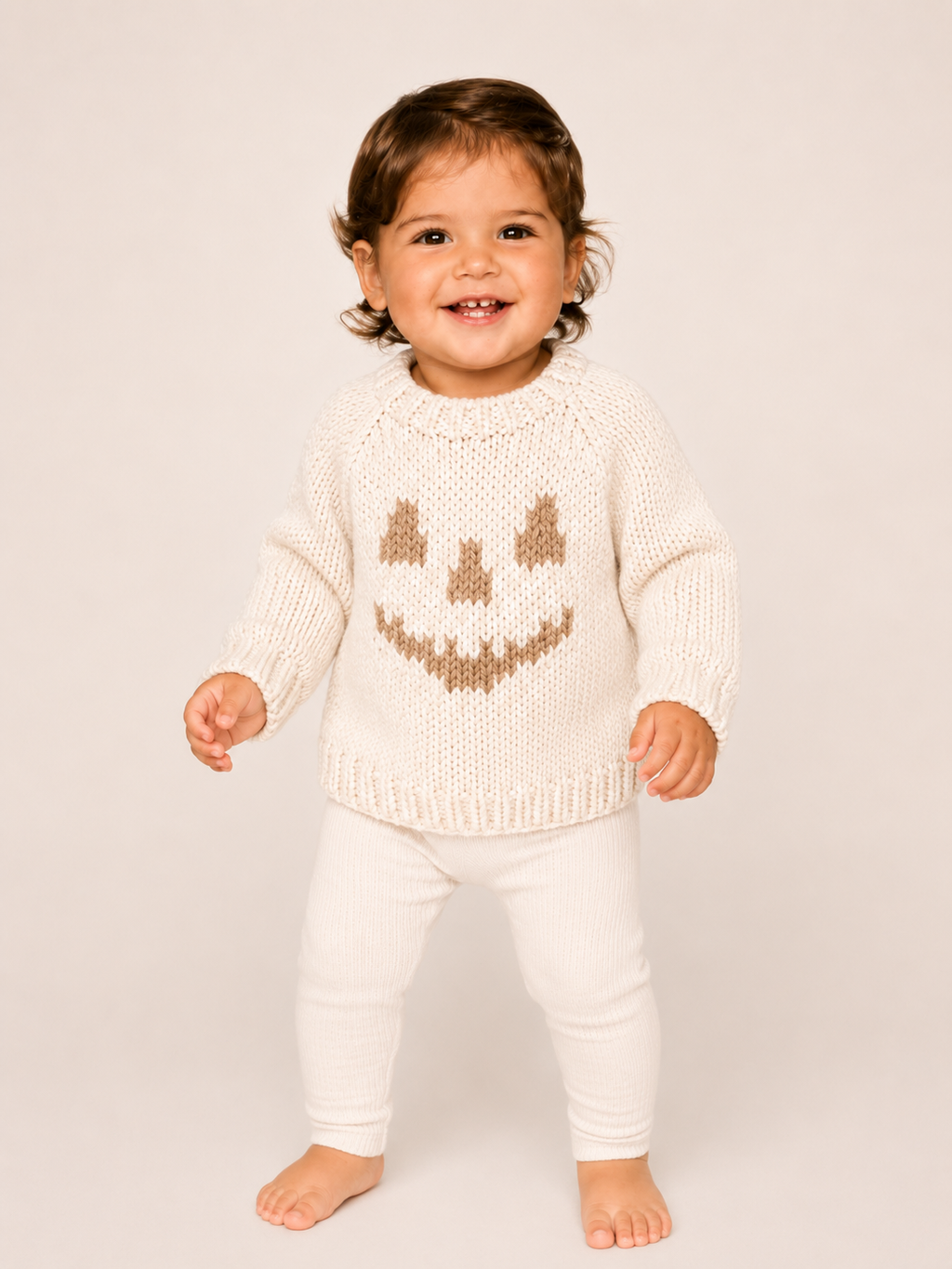 A smiling toddler standing barefoot against a plain light beige studio background, wearing a cream knit sweater with a brown knitted jack-o'-lantern face and matching cream pants.