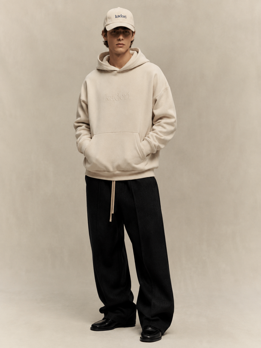 A model stands against a neutral beige studio backdrop wearing a cream-colored embroidered hoodie, black wide-leg pants, black shoes, and a matching cream cap with 'kith' embroidered on it.