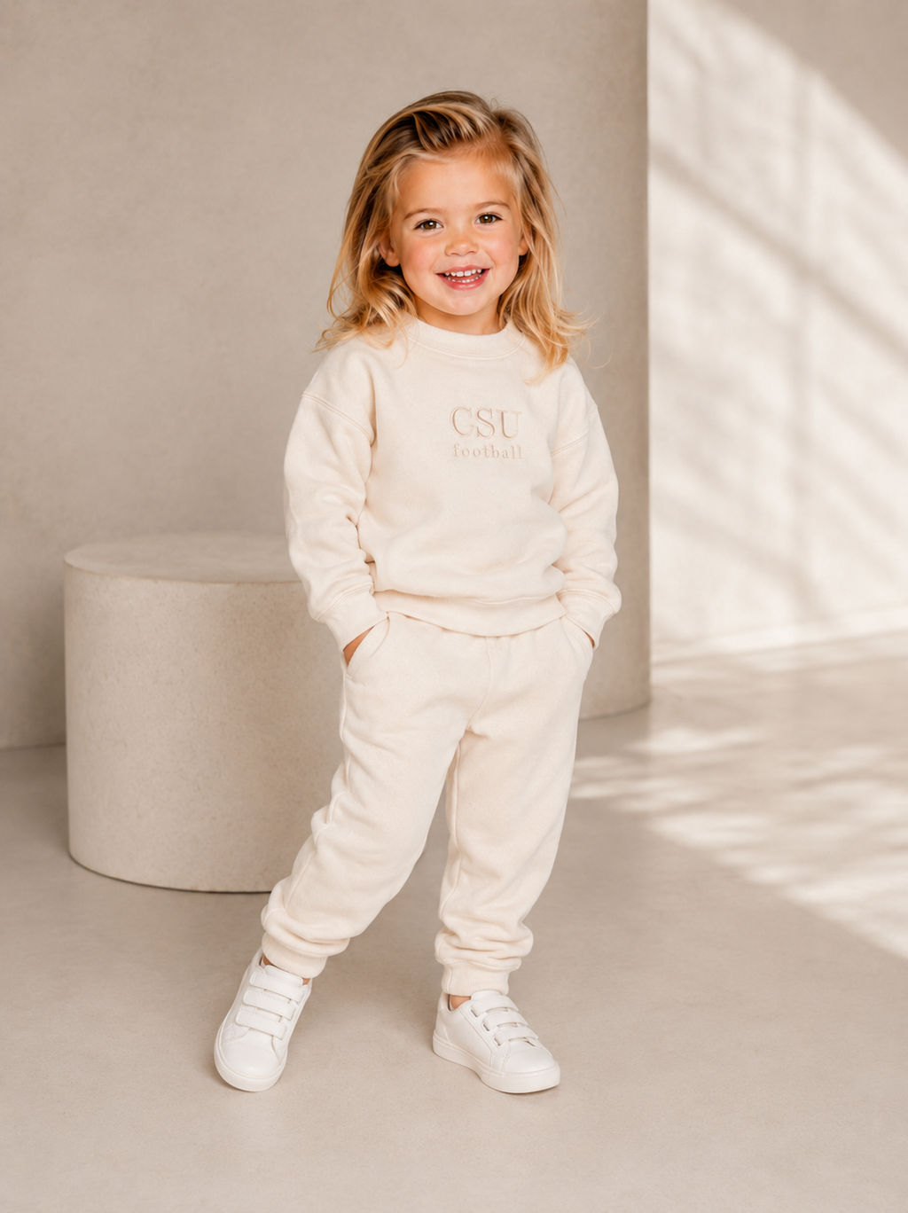 A young girl smiling and posing in a matching cream-colored sweatshirt and jogger pants set, with 'CSU Coachella' printed on the chest, standing in a minimalist studio setting.