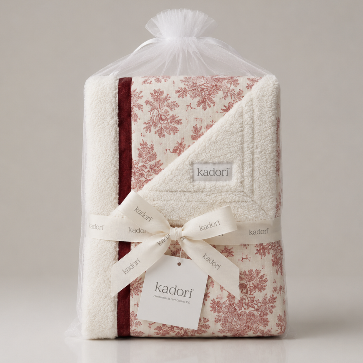 A folded Kadori baby blanket wrapped in a clear tulle bag, tied with a cream ribbon and bow. The blanket features a cream background with a dusty rose floral toile pattern and a dark red border stripe.