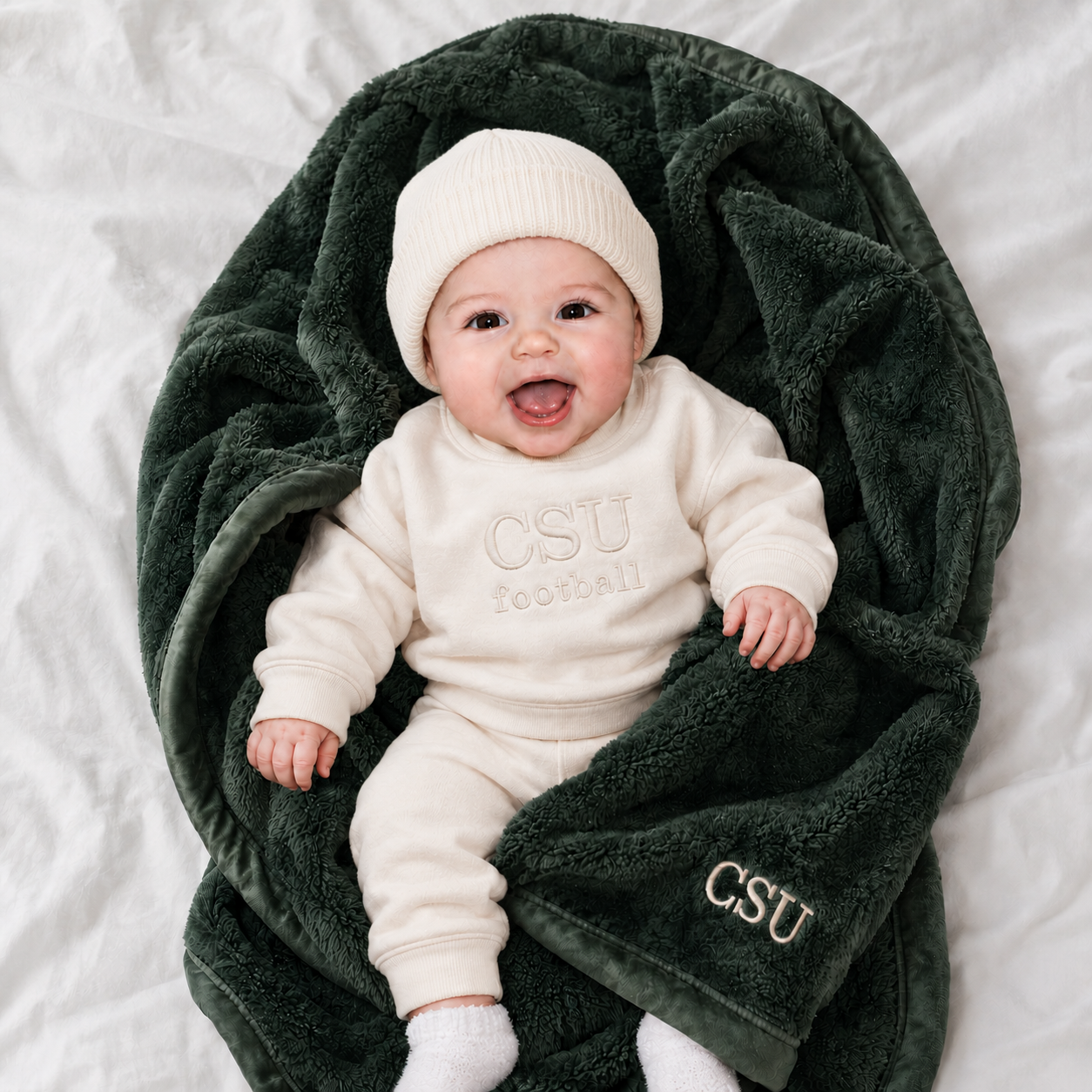 A baby lying on a white sheet, wrapped in a dark forest green plush blanket with 'CSU' embroidered in gold on the corner. The baby is wearing a cream-colored sweatshirt and pants with 'CSU football' embroidered on the chest, along with a cream knit beanie and white socks.