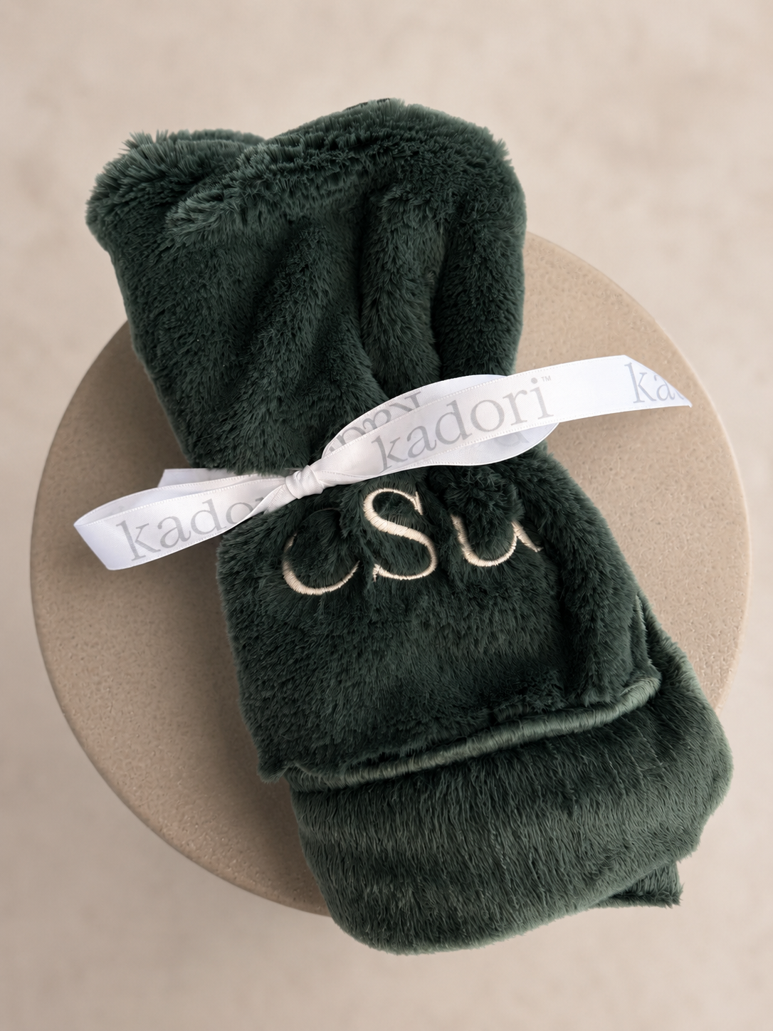 A dark forest green plush bath towel folded and tied with a white ribbon printed with the brand name 'kadori', featuring gold embroidered initials 'CSU' on the front, displayed on a round beige surface.