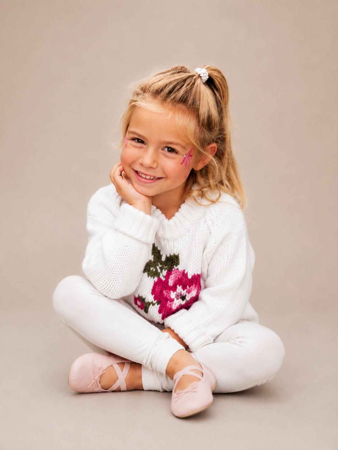 A young girl with blonde hair in a ponytail sits cross-legged on a beige studio background, wearing a white knit sweater with a pink and green floral design, white leggings, and pink ballet slippers.
