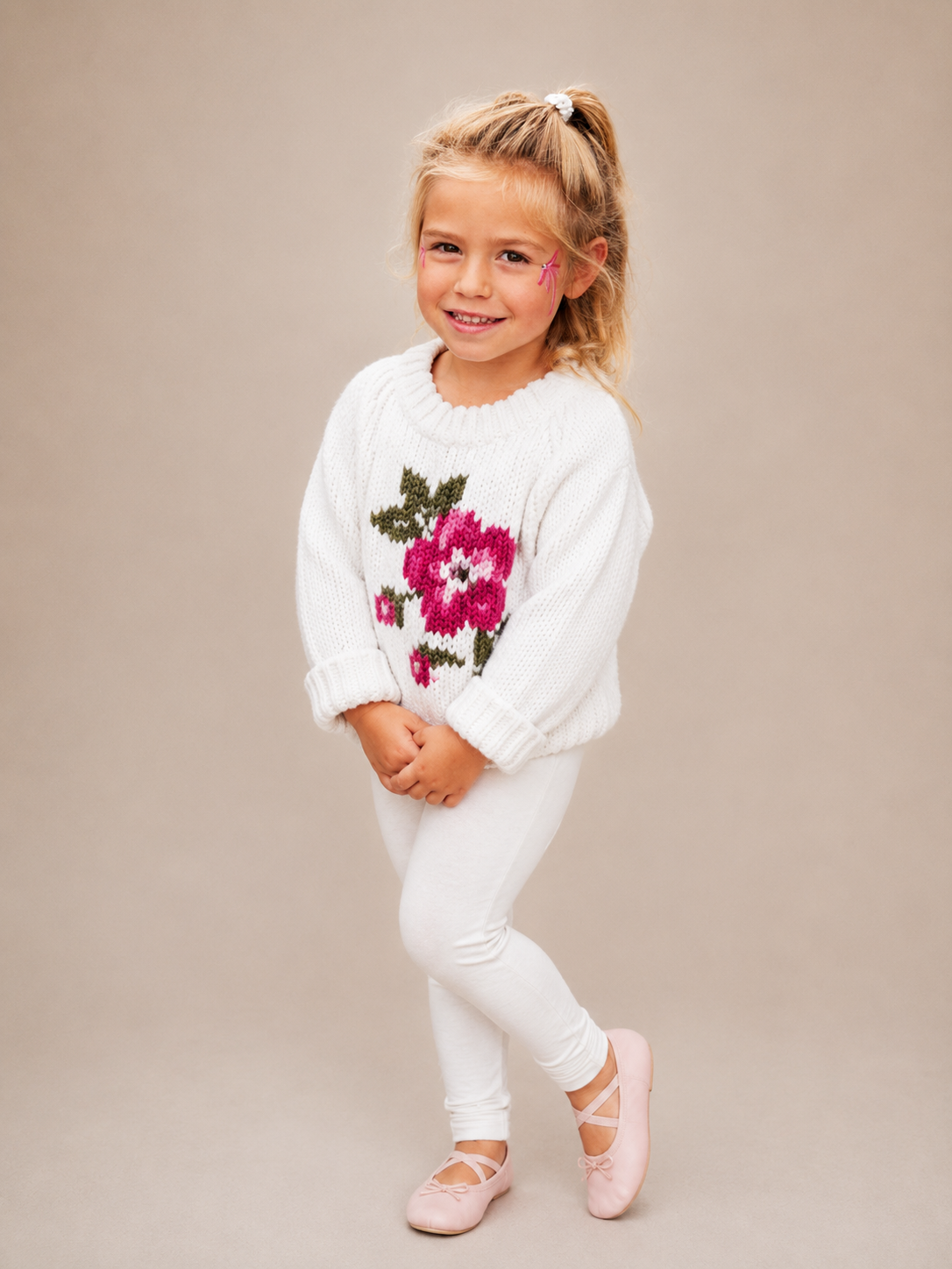 A young girl with blonde hair in a ponytail stands against a solid beige studio background, wearing a white knit sweater with a large pink and green floral pattern on the front, white leggings, and light pink ballet flats.