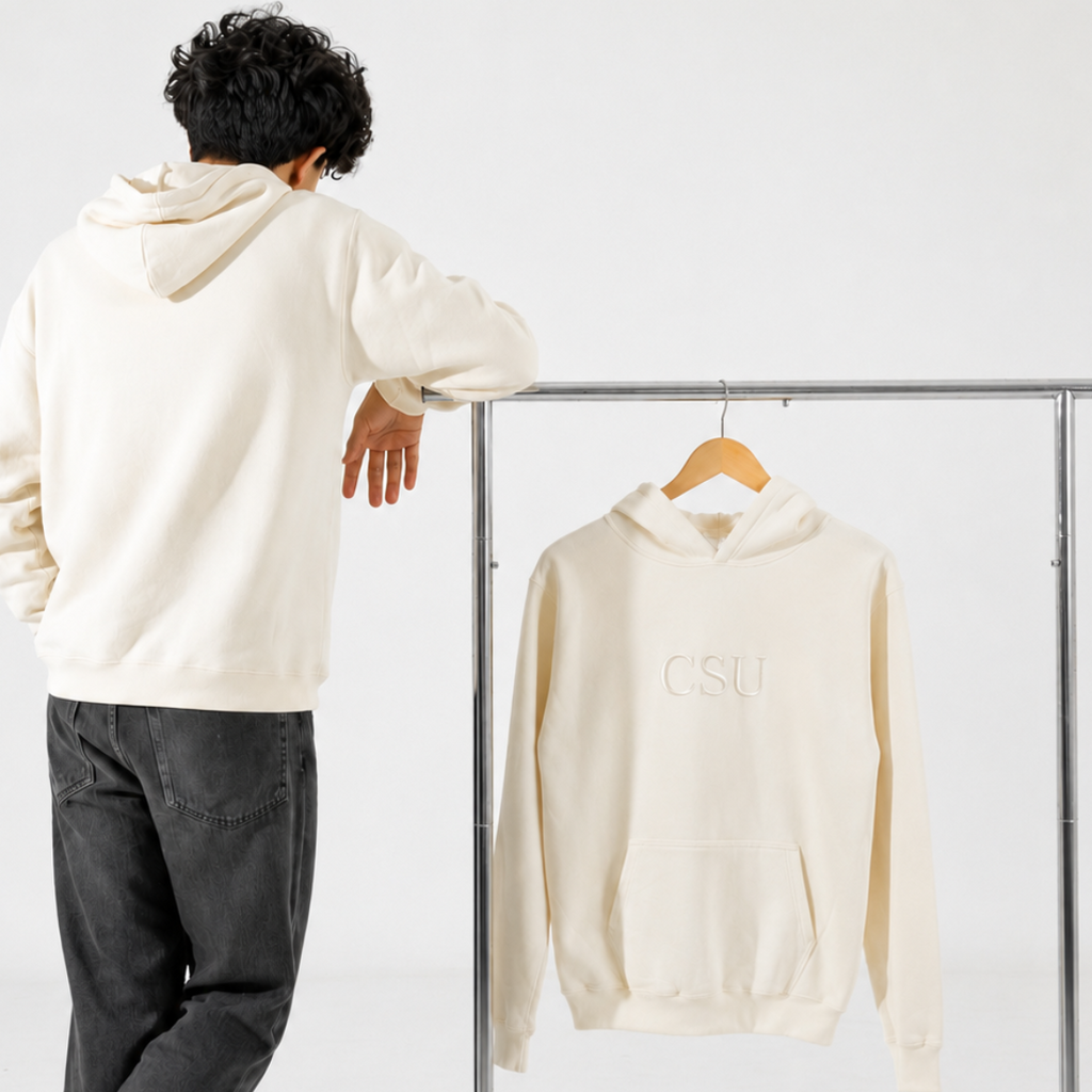 A person stands with their back to the camera next to a metal clothing rack, wearing a cream hoodie and dark gray jeans. The cream hoodie with a subtle 'CSU' logo on the chest is hanging on a wooden hanger on the rack.