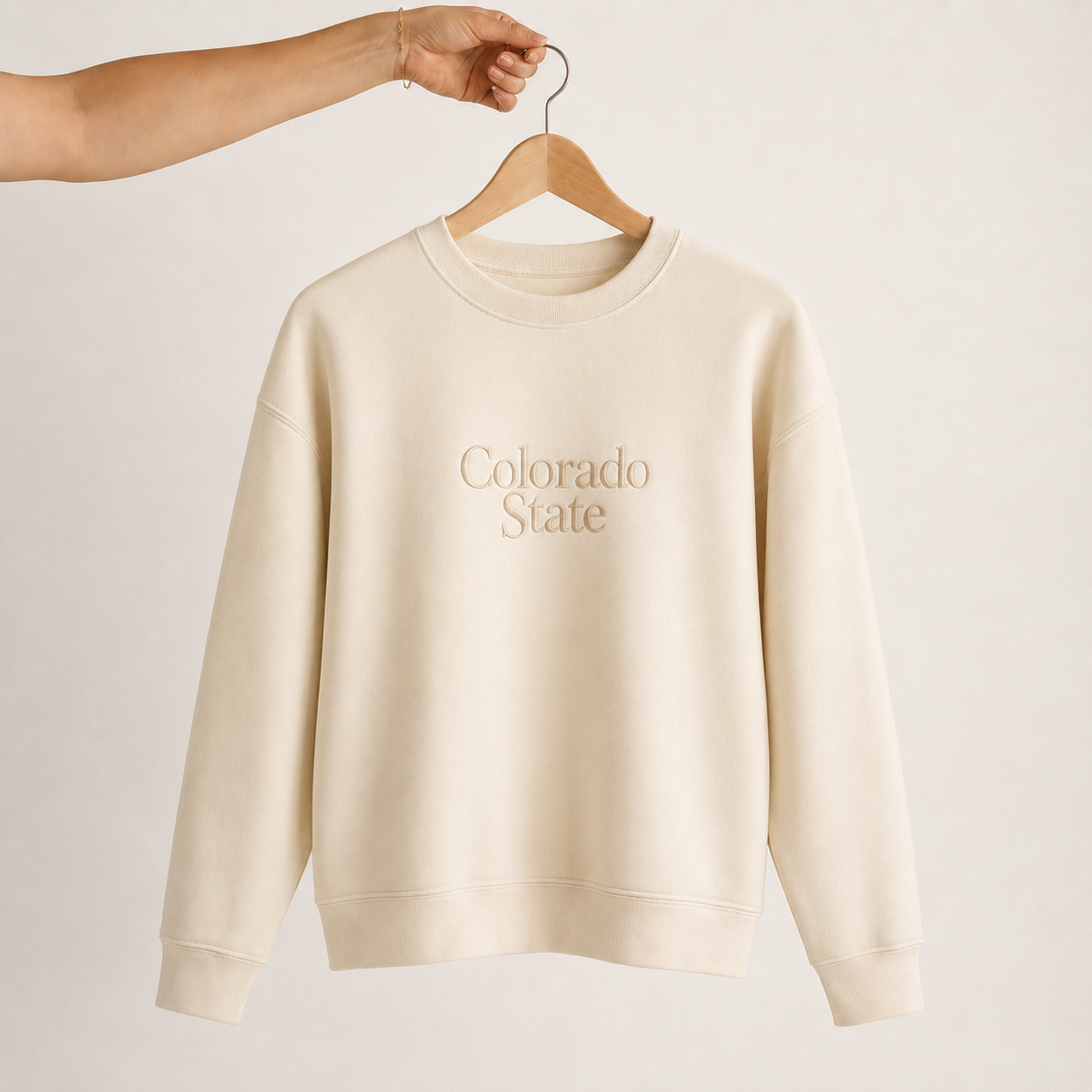 An off-white crewneck sweatshirt with 'Colorado State' printed in a matching beige font on the chest, hanging on a wooden hanger held by a person's arm against a light beige background.