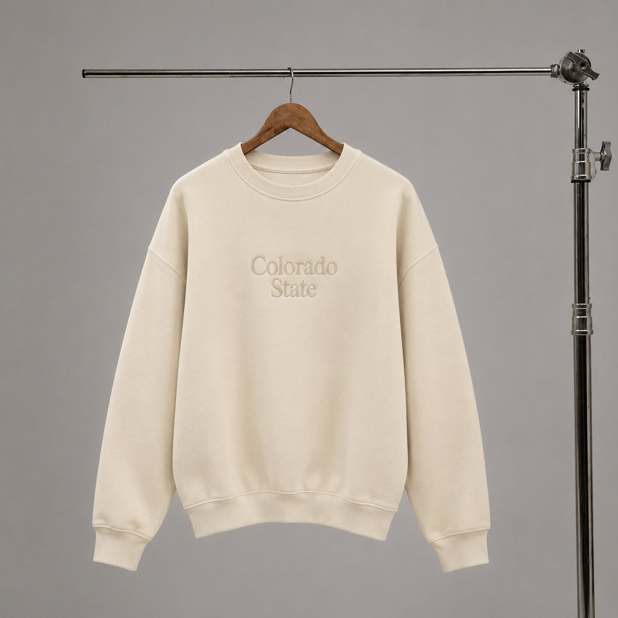An off-white crewneck sweatshirt with 'Colorado State' embroidered in beige on the chest, hanging on a wooden hanger from a metal clothing rack against a solid gray background.