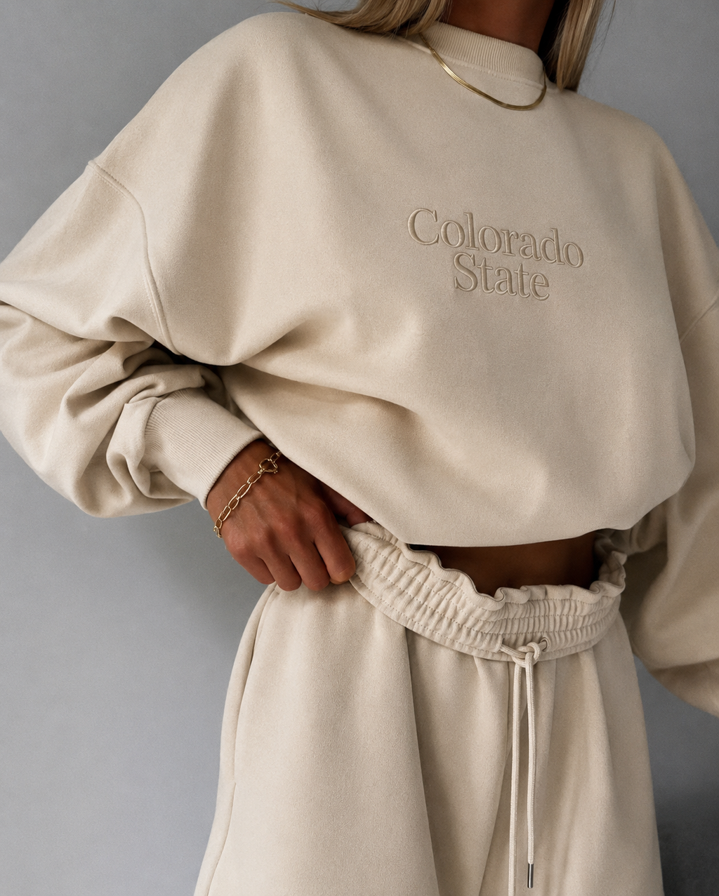 A person wearing a cream-colored sweatshirt with 'Colorado State' embroidered on the chest, paired with matching sweatpants and gold jewelry, against a neutral gray background.