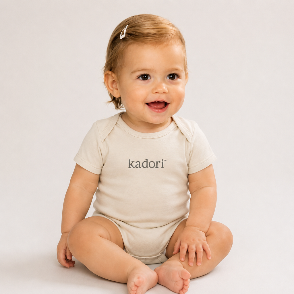 A baby sitting cross-legged wearing a short-sleeve cream bodysuit with the brand name 'kadori' printed in black across the chest, set against a plain light gray background.