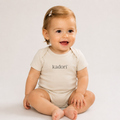 A baby sitting cross-legged wearing a short-sleeve cream bodysuit with the brand name 'kadori' printed in black across the chest, set against a plain light gray background.