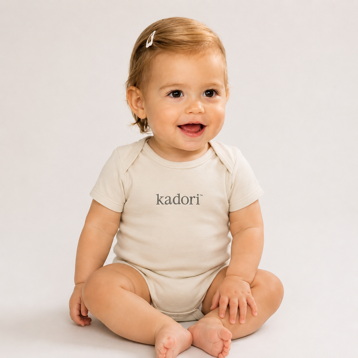 A baby sitting cross-legged wearing a short-sleeve cream bodysuit with the brand name 'kadori' printed in black across the chest, set against a plain light gray background.