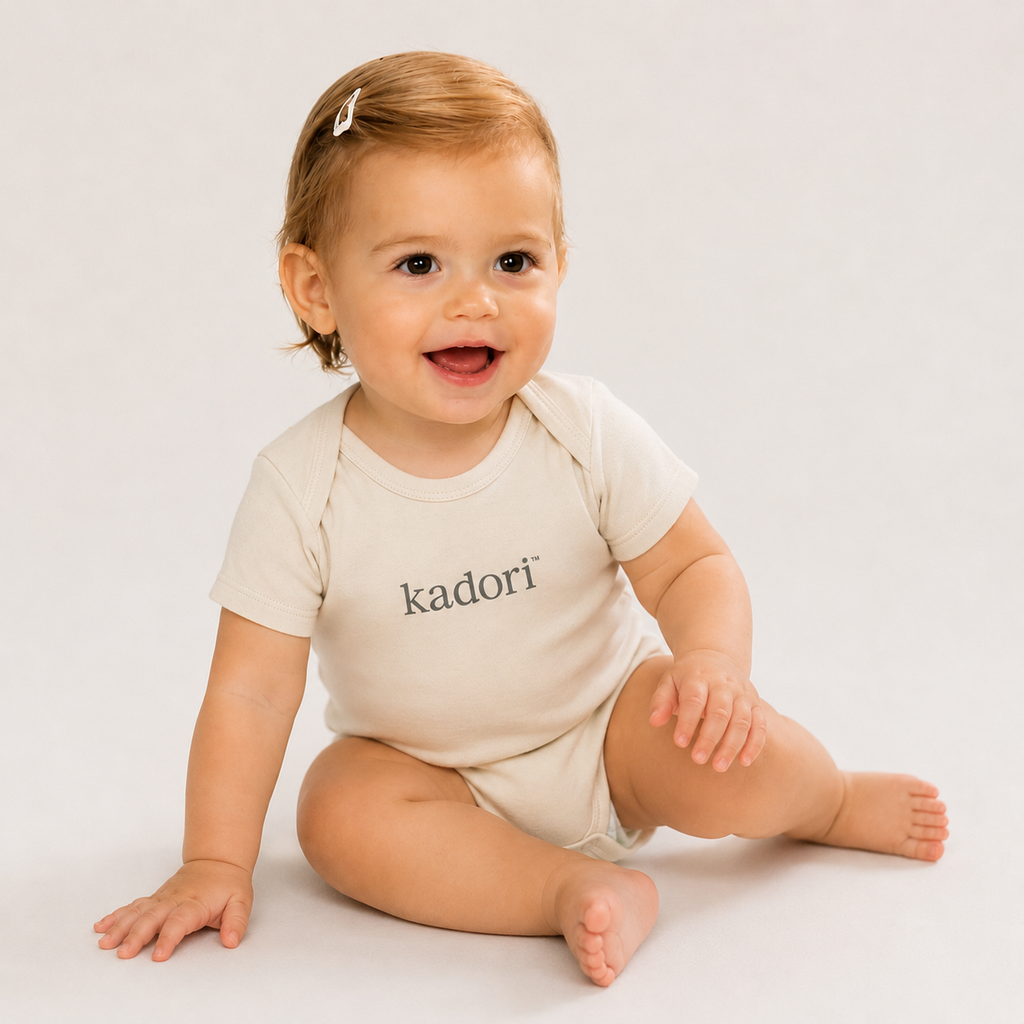 A baby sitting on a light studio background wearing a cream short-sleeve bodysuit with the name 'kadori' printed in black across the chest.