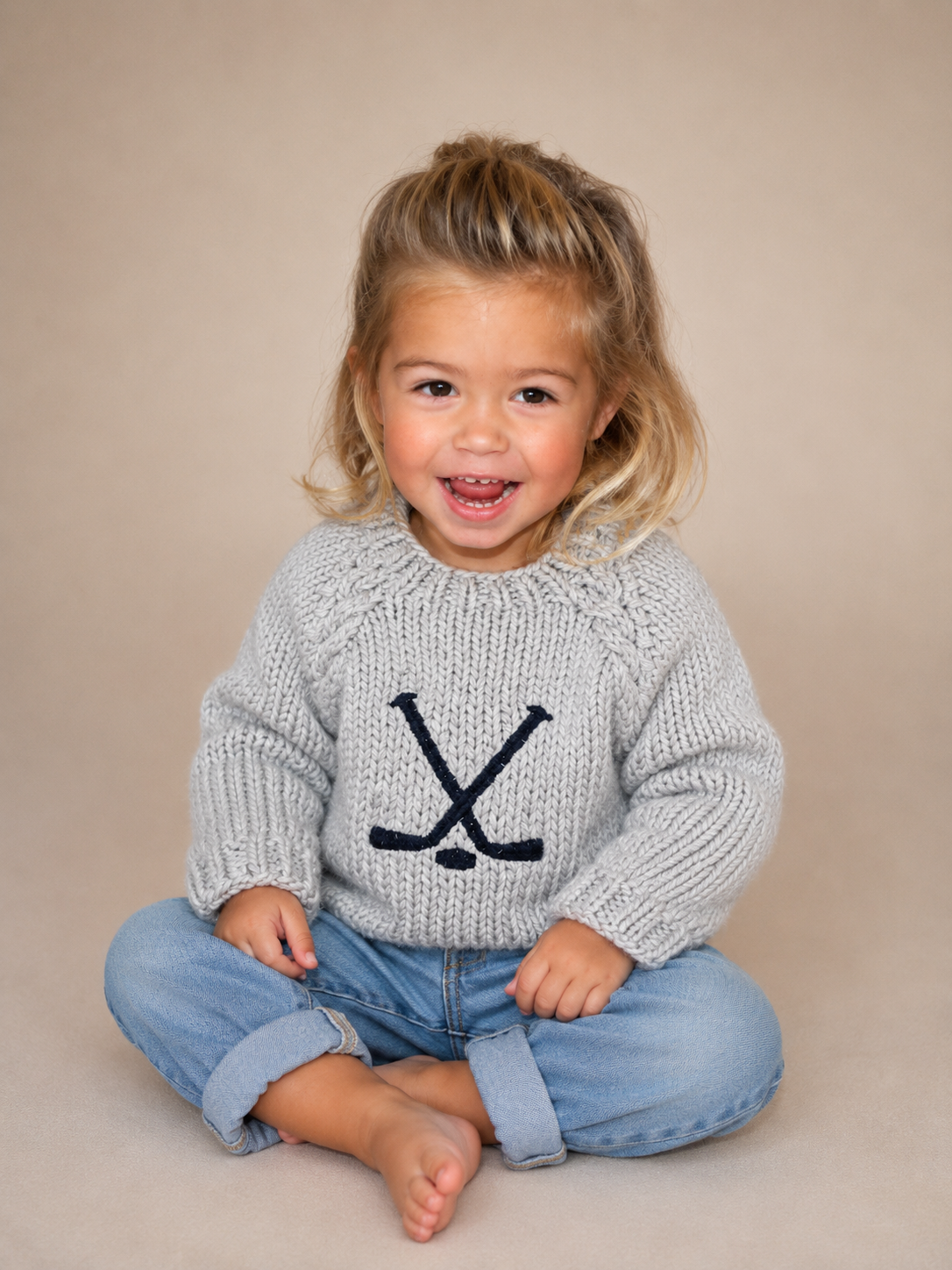 A young child sitting cross-legged on a beige studio background, wearing a light gray knit sweater with a dark navy embroidered hockey stick and letter design on the chest, paired with light blue jeans.