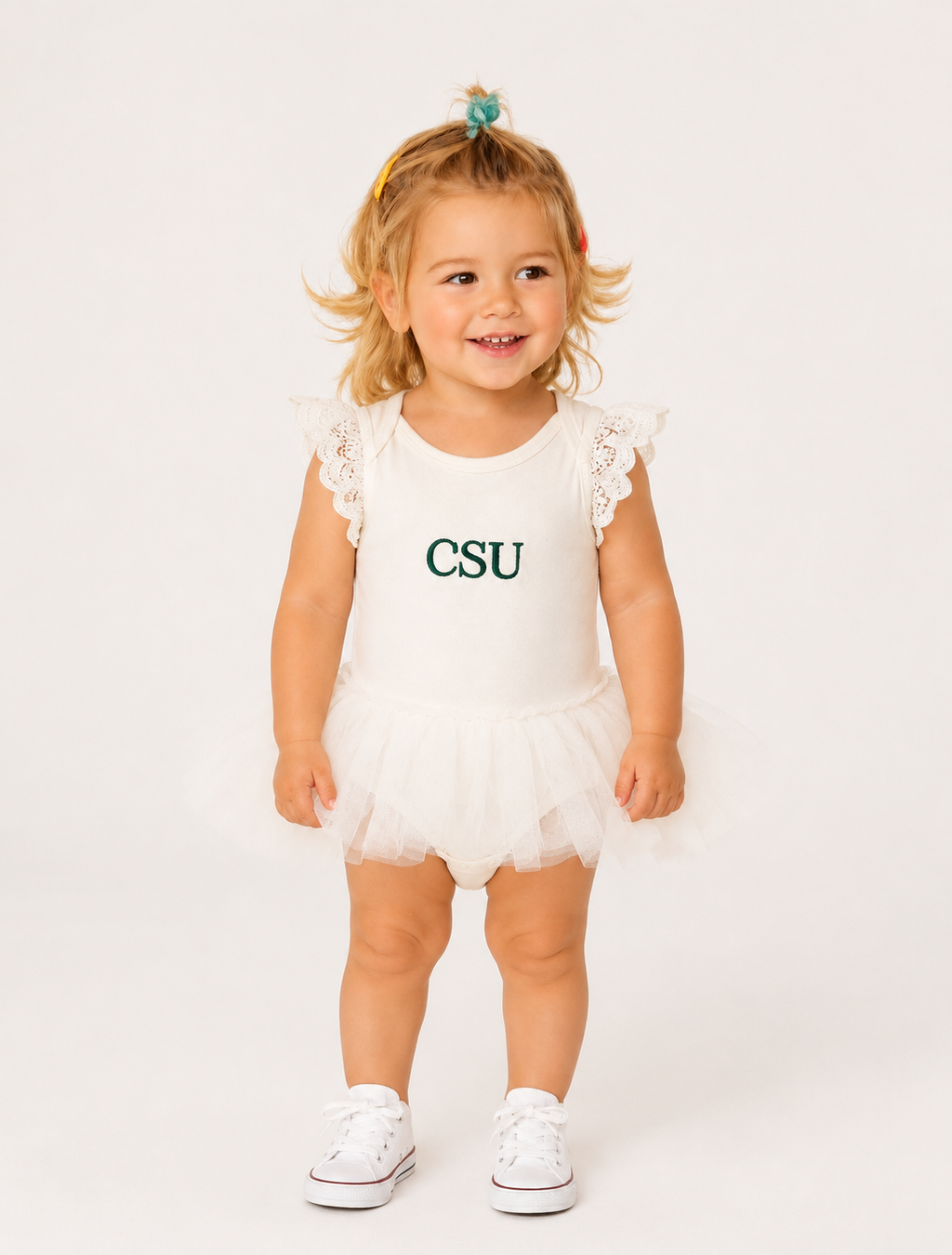 A toddler girl with blonde hair in a small blue bow stands against a white studio background wearing a white sleeveless tutu romper with lace flutter sleeves and 'CSU' printed in dark green on the chest, paired with white sneakers.