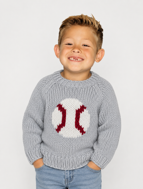 A young boy smiling while wearing a light gray knit sweater with a baseball design on the front, paired with blue jeans, against a plain light gray background.