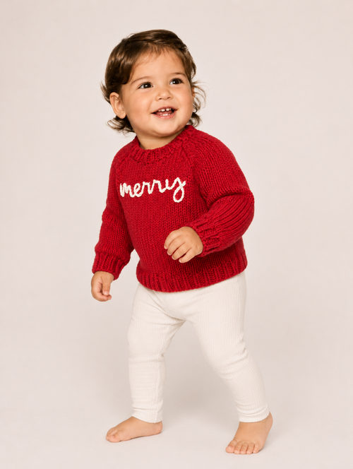 A toddler standing barefoot against a light beige studio background, wearing a red knit sweater with 'merry' in white script across the chest and white pants.