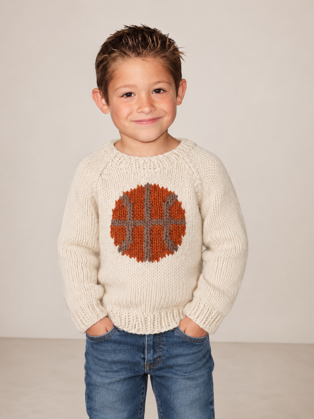 A young boy wearing a cream-colored knit sweater with a large orange and gray basketball motif on the chest, paired with blue jeans, standing against a neutral beige background.