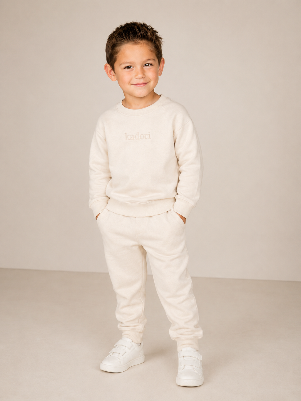 A young boy modeling a cream-colored two-piece loungewear set consisting of a sweatshirt and sweatpants, paired with white sneakers, against a neutral beige studio background.