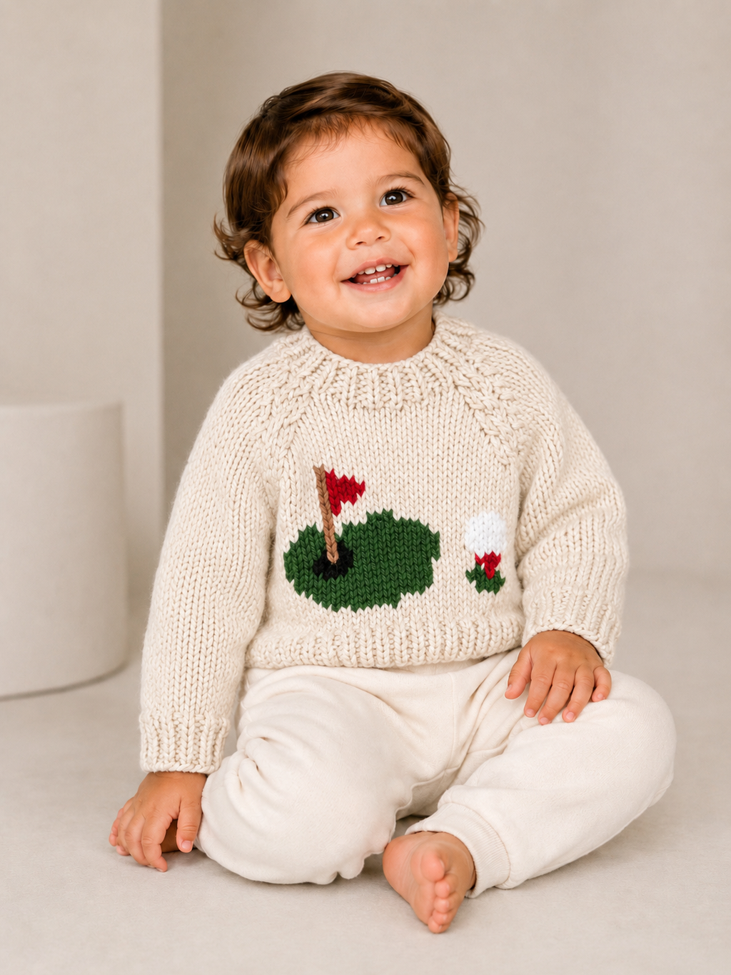 A smiling toddler sitting on a light beige floor wearing a cream-colored knit sweater with a knitted green lily pond and red flower design on the front, paired with cream pants.