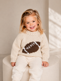 A young child wearing a cream knit sweater with a brown and white football graphic on the chest, paired with cream pants, sitting on a round beige stool.