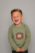 A young boy with short blond hair smiling and laughing, wearing a sage green knit sweater with a brown and white football graphic on the chest, set against a solid gray background.