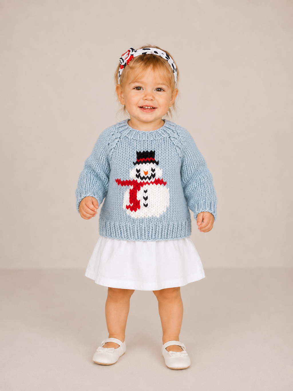 A toddler standing against a plain beige studio background, wearing a light blue knit sweater with a snowman motif, a white skirt, white shoes, and a patterned headband.
