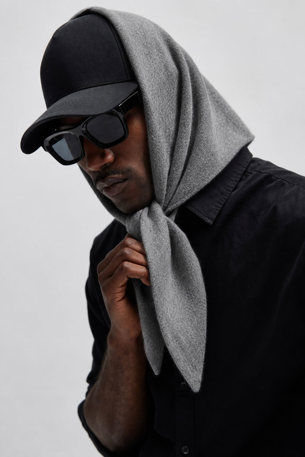A man wearing a black baseball cap, black sunglasses, and a black button-up shirt, with a light gray knit hood draped over his head and neck, photographed against a plain light gray background.