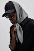 A man wearing a black baseball cap, black sunglasses, and a black button-up shirt, with a light gray knit hood draped over his head and neck, photographed against a plain light gray background.