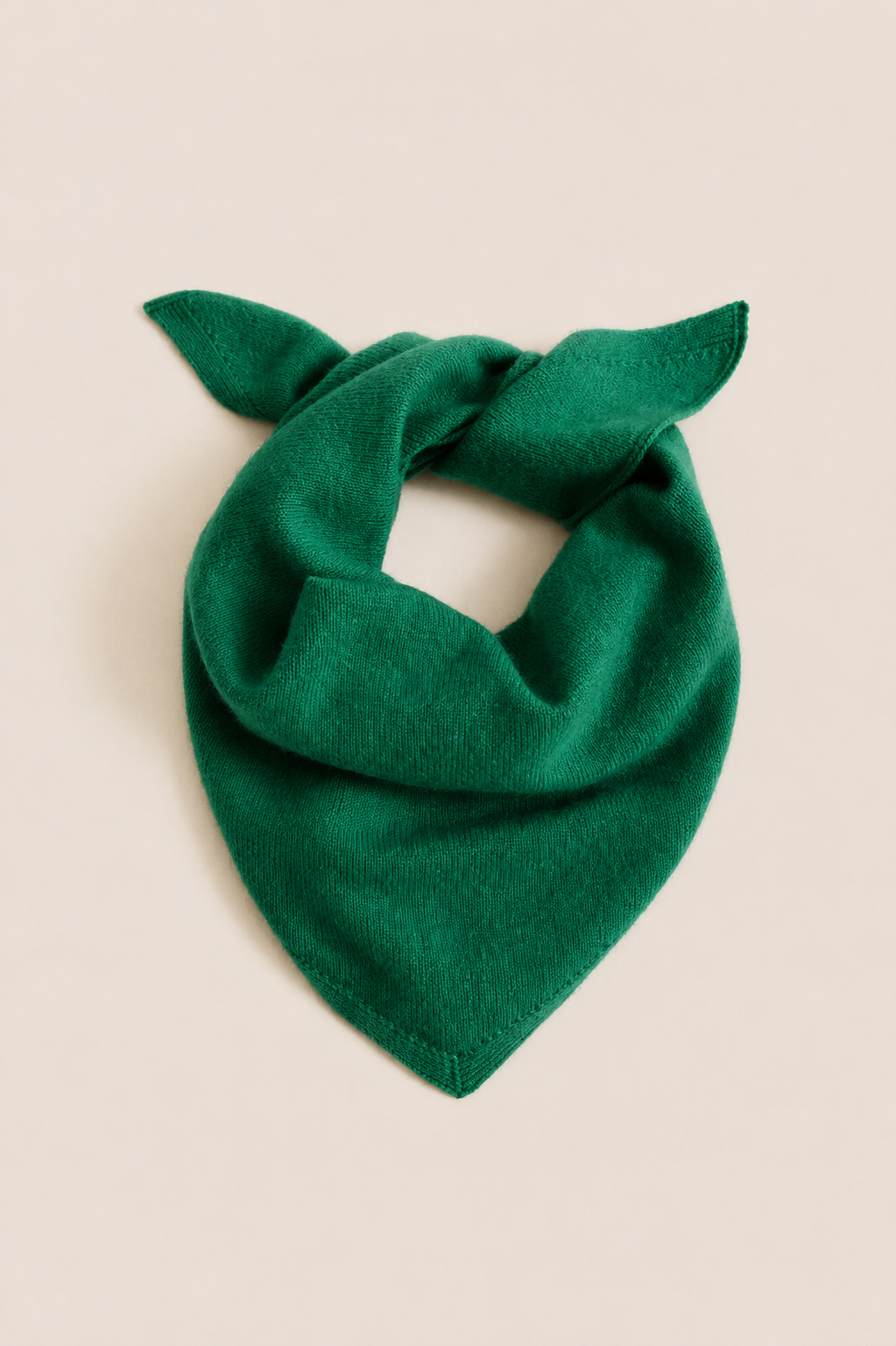 A dark green knit bandana or neck warmer scarf with two small looped ends, styled in a knot on a solid light beige background.