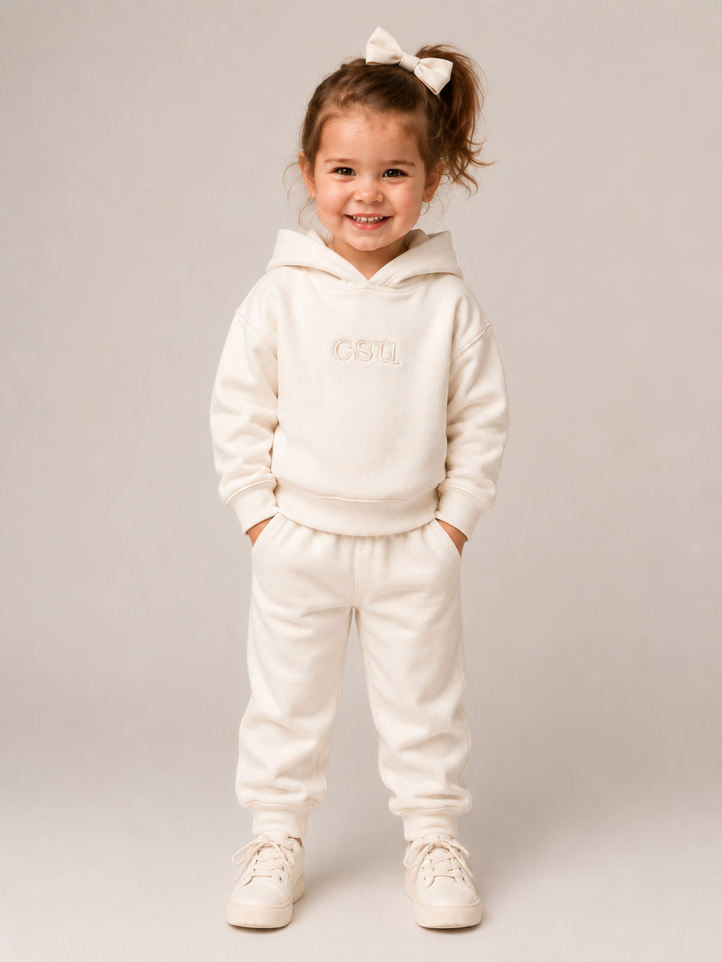 A young girl with a white bow in her hair stands smiling, wearing a matching cream-colored two-piece loungewear set consisting of a hooded sweatshirt with 'CSTL' embroidered on the chest and matching jogger pants, paired with cream sneakers against a neutral studio background.