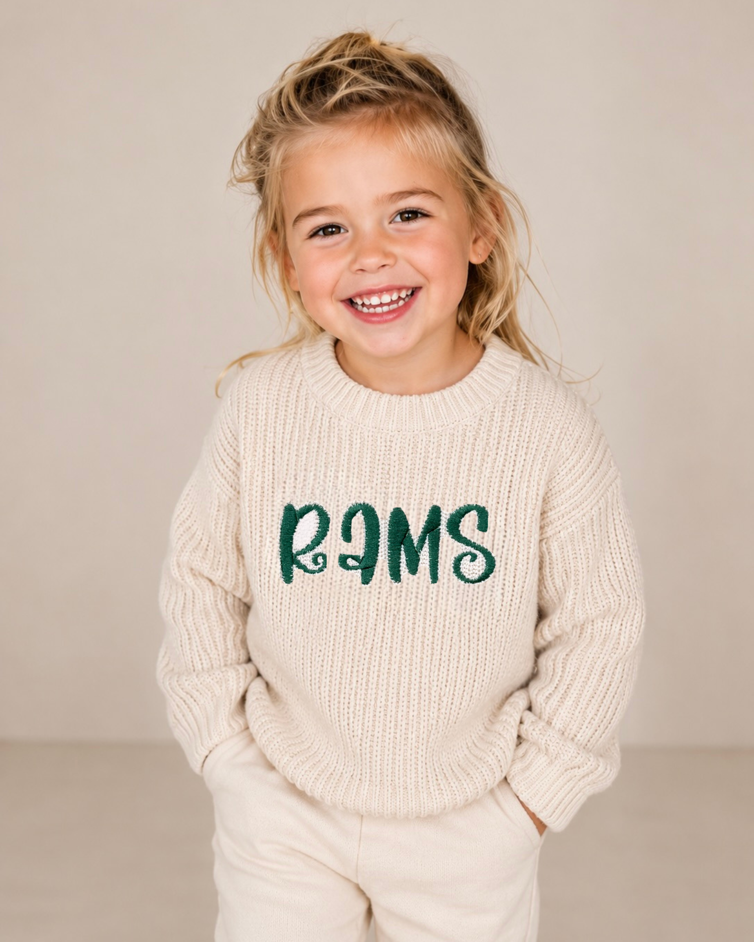 A young girl smiling and wearing a cream-colored ribbed knit sweater with the word 'RAMS' in dark green across the chest, paired with matching cream pants, set against a plain beige studio background.