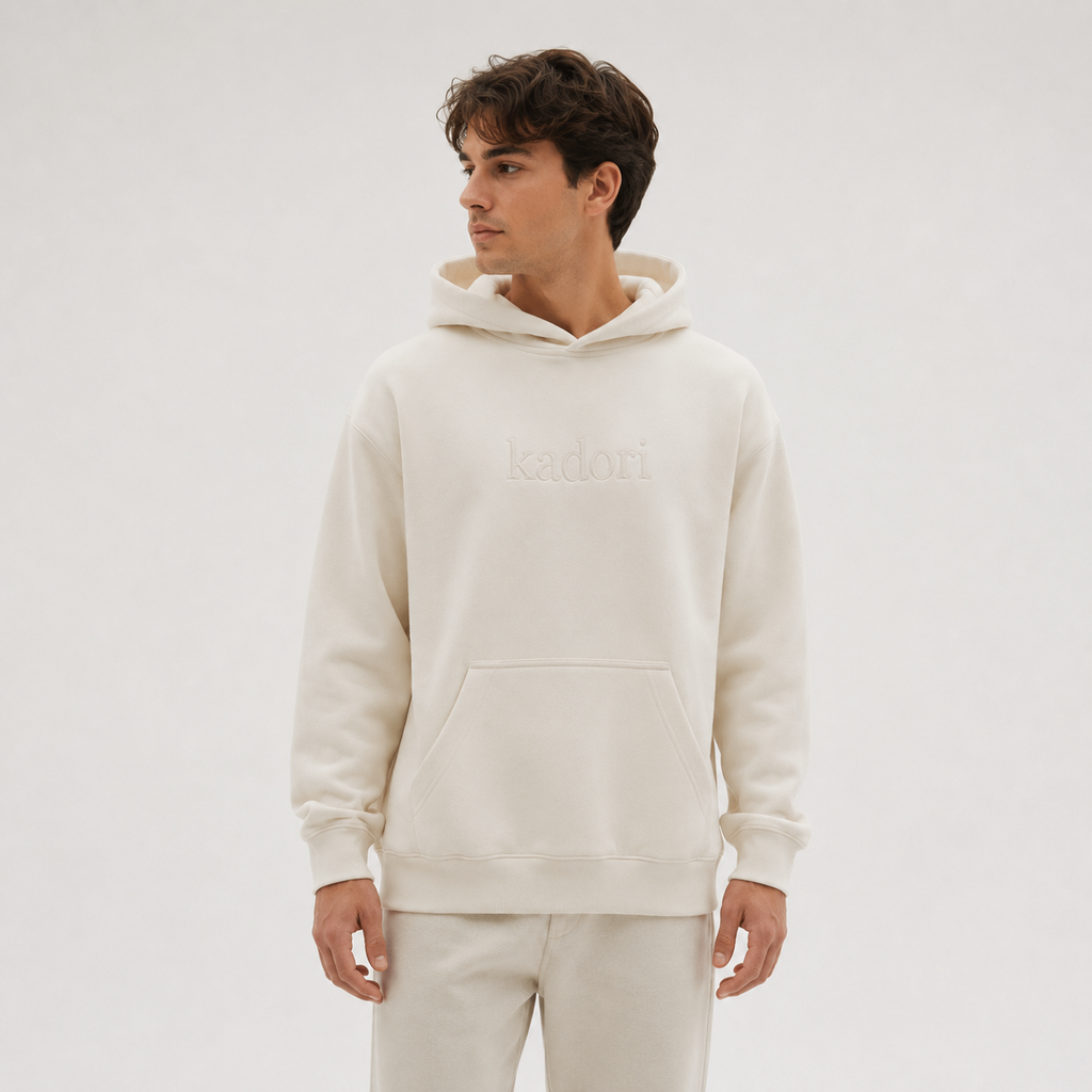 A man wearing a cream-colored hoodie with a tonal embroidered 'kadori' logo on the chest, paired with matching cream sweatpants, set against a plain light gray studio background.