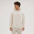 A man wearing a cream-colored hoodie with a tonal embroidered 'kadori' logo on the chest, paired with matching cream sweatpants, set against a plain light gray studio background.