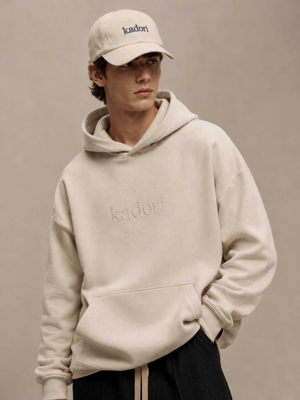 A model wearing a beige Kadori embroidered hoodie and matching beige baseball cap, posed against a neutral beige studio background.
